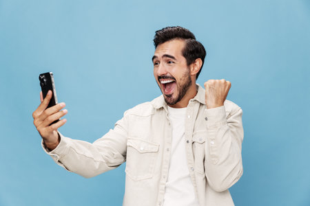 Portrait of a brunette man with a phone in his hands, a smile with teeth of surprise and happiness, on a blue background in a white T-shirt and jeans, copy spaceの写真素材