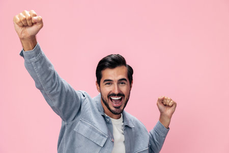 Fashion portrait of a brunette man with a beard happiness victory raised his hands with his fist up on a pink background in a white T-shirt smile and joyful emotion on his face, copy spaceの写真素材