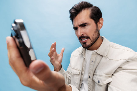 Portrait of a stylish brunette man looking at the phone blogger with a beard of anger and frustration, losing the game on a blue background in a white T-shirt and jeans, copy spaceの写真素材