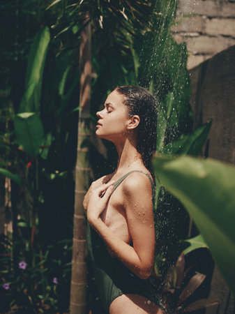 Sexy woman, body in a swimsuit takes a tropical shower outdoors against the backdrop of green tropical leaves, flowers and palm trees. Body and hair care, tanned skin, sunlight, smile, vintageの写真素材