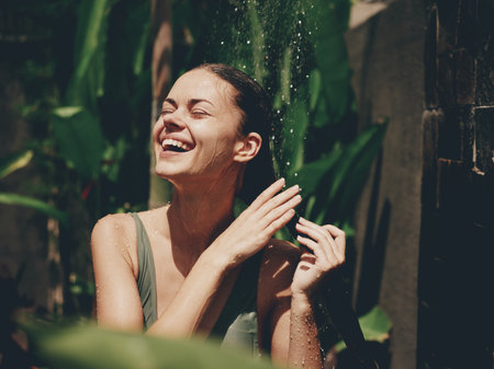 A woman, a body in a swimsuit washes her head in a tropical shower outdoors against backdrop green tropical leaves, flowers and palm trees. Body and hair care, tanned skin, sunlight, smile, vintageの写真素材