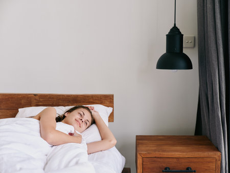 Woman in the morning lying on the bed under the blanket in the bedroom, morning awakening and stretching her arms, smiling on a day off, lifestyleの写真素材