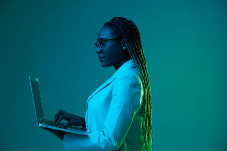 African African woman with laptop in glasses on a blue background in neon light, color mixed light. Data storage cyber security, hacker, big data.の写真素材
