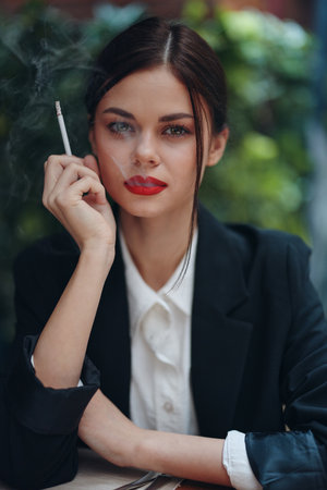 Stylish fashion woman portrait sitting in a cafe at a table and smoking a cigarette releasing smoke from her mouth with red lipstick, a bad habit, a smile with teeth and a thoughtful lookの写真素材