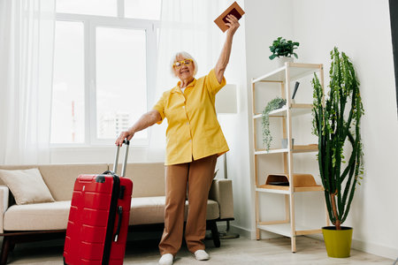 Happy senior woman with passport and travel ticket packed a red suitcase, vacation and health care. Smiling old woman joyfully stands in the house before the trip.の写真素材