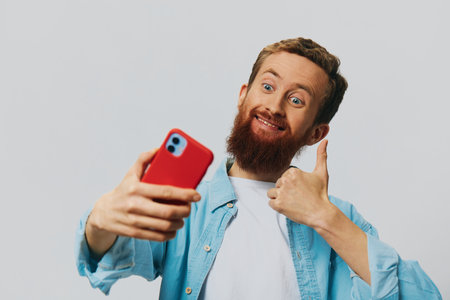 Man hipster with phone in hand blogger smile on gray background in blue shirt and white t-shirt, talking on the phone and selfiesの写真素材