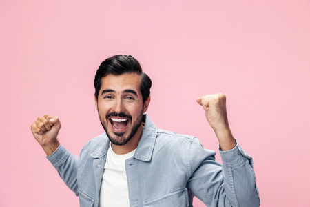 Fashion portrait of a brunette man with a beard happiness victory raised his hands with his fist up on a pink background in a white T-shirt smile and joyful emotion on his face, copy spaceの写真素材