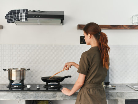 A woman at home smile in the kitchen prepares food stands with her back to the camera at the stove and fries meat and vegetables in a pan, a large kitchen in a modern style, lifestyleの写真素材
