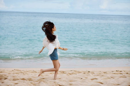 Sports woman runs along the beach in summer clothes on the sand in a yellow tank top and denim shorts white shirt flying hair ocean view, beach vacation and travelの写真素材