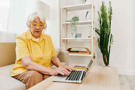 Happy elderly woman with a laptop typing with headphones sitting at home on the couch in a yellow shirt, bright modern interior, lifestyle online communication.の写真素材