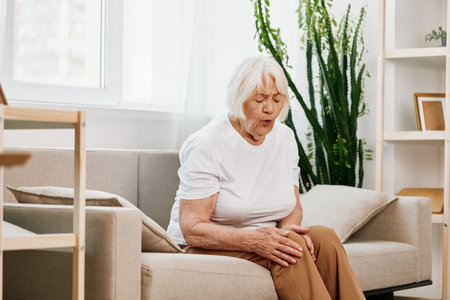 Elderly woman severe pain in her leg sitting on the couch, health problems in old age, poor quality of life. Grandmother with gray hair holds on to her sore knee, problems with joints and ligamentsの写真素材