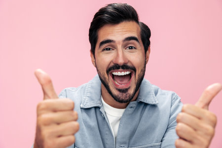 Fashion portrait of a brunette man with a beard showing a thumbs up sign on a pink background in a...