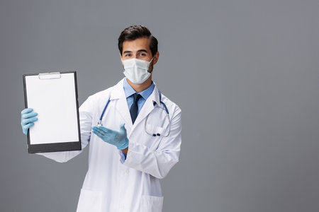 A male doctor in a white coat and a medical mask with a notepad in his hand looks at the camera on a gray isolated background, copy space, space for textの写真素材