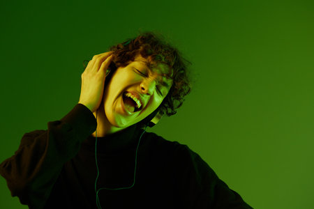 Man wearing headphones listening to music, dancing and singing with his eyes closed, DJ happiness and smile, hipster lifestyle, portrait green background mixed neon light, copy spaceの写真素材