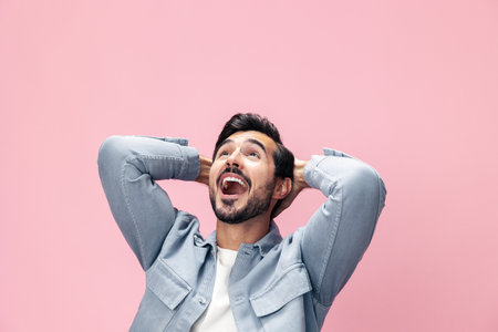 Fashion portrait of a brunette man with a beard happiness victory raised his hands with his fist up on a pink background in a white T-shirt smile and joyful emotion on his face, copy spaceの写真素材