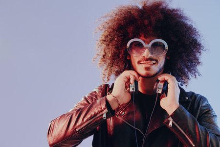 Portrait of a stylish man with curly hair with glasses and headphones on a blue background multinational, colored light, black leather jacket trend, modern concept.の写真素材