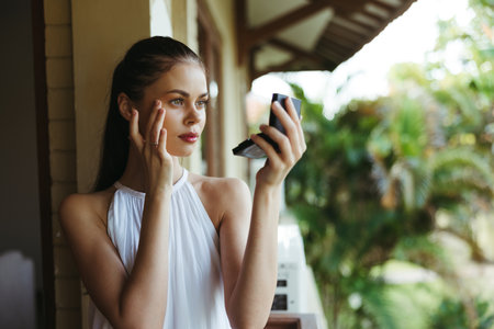 A young beautiful woman looks at her face with makeup in the mirror outside in the summer, protective spf products for problem skin with acne.の写真素材