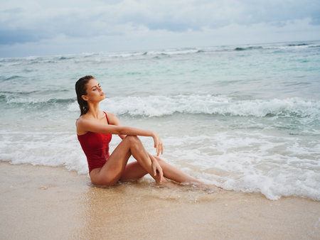 A young woman in a red swimsuit sits on the sand with a beautiful sun tan and looks out at the ocean in the tropics on an island beachの写真素材
