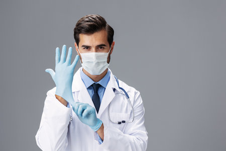 A male doctor in a white coat and medical mask and sterile gloves looks at the camera on a gray isolated background, copy space, space for textの写真素材