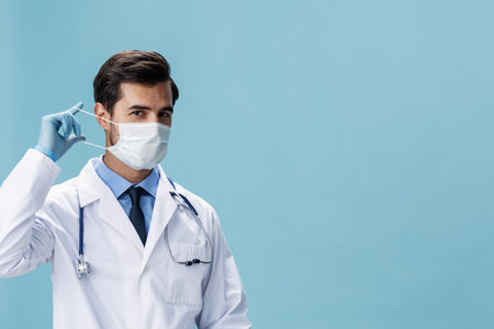 Male doctor in a white coat and medical mask looks at the camera on a blue isolated background, copy space, space for textの写真素材
