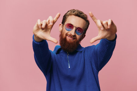 Portrait of a redheaded man wearing sunglasses smiling and dancing, listening to music on a pink background. Hipster with a beard, happiness finger pointing.の写真素材