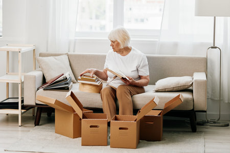 elderly woman sits on a sofa at home with boxes. collecting things with memories albums with photos and photo frames moving to a new place cleaning things and a happy smile. Lifestyle retirement.の写真素材