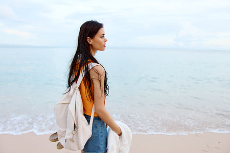 A young woman after swimming in the ocean with a backpack in wet clothes walks along the beach, summer vacation on an island by the ocean in Bali sunsetの写真素材