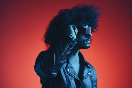 Portrait of fashion man with curly hair on red background with stylish glasses, multicultural, colored light, black leather jacket trend, modern concept.の写真素材