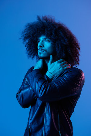 Portrait of fashion man with curly hair on blue background multinational, colored light, black leather jacket trend, modern concept.の写真素材