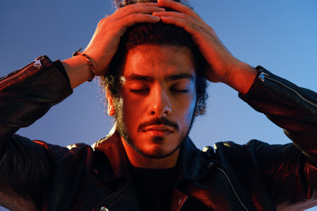 Portrait of a stylish man with curly hair on a blue background multinational, colored light, black leather jacket trend, modern concept.の写真素材
