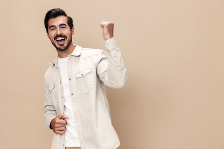 Portrait of a stylish man smile with teeth and surprise raised his hands with fists up on a beige background in a white t-shirt, fashionable clothing style, copy space, space for textの写真素材