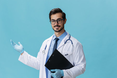 Male doctor in a white coat and glasses with a medical record and a stethoscope smile on a blue isolated background, copy space, space for textの写真素材