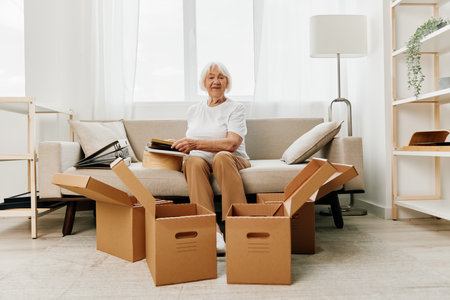 elderly woman sits on a sofa at home with boxes. collecting things with memories and moving and happiness smile.の写真素材