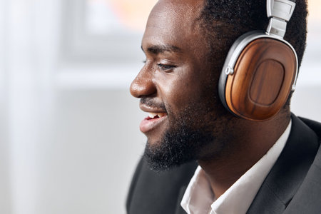 An African-American man sits at his desk in front of his laptop, wearing headphones and chatting on...