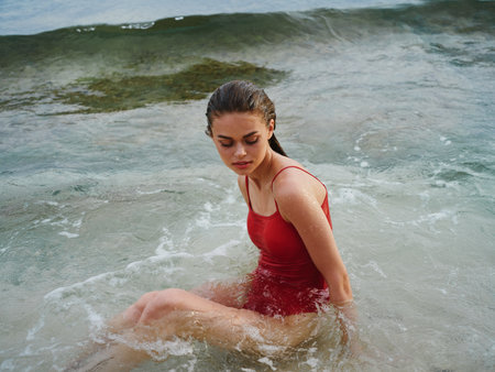 A happy woman in a swimsuit with wet hair sits in the ocean in the waves on a surfing beach and looks into the camera, a strong look, sunset on a tropical islandの写真素材
