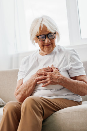 Elderly woman severe chest pain sitting on the sofa, health problems in old age, poor quality of life. Grandmother with gray hair holds her chest with her hands, womens health, breast cancer.の写真素材