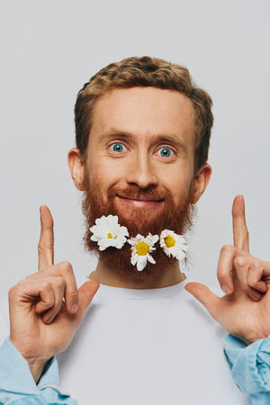 Portrait of a funny man in a white T-shirt with flowers daisies in his beard on a white isolated background, copy place. Holiday concept and congratulations.の写真素材