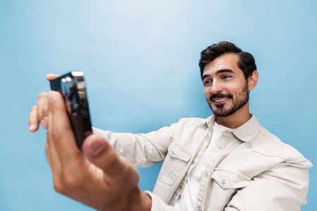 Portrait of a stylish brunette man shooting himself on the phone blogger with a beard beautiful smile on a blue background in a white T-shirt and jeans smile and joyful emotion on his face, copy spaceの写真素材