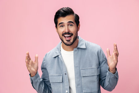 Portrait of a stylish brunette man with a beard raised his hands up with surprise open pink background in a white T-shirt and jeans smile and joyful emotion on his face, copy spaceの写真素材