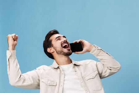 Portrait of a man brunette animation and joy talking on the phone hand and fist up from victory joy, on a blue background in a white T-shirt and jeans, copy spaceの写真素材