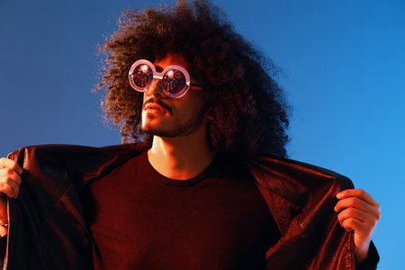 Portrait of a stylish man with curly hair with glasses smile on blue background multinational, color light, black leather jacket trend, modern concept.の写真素材