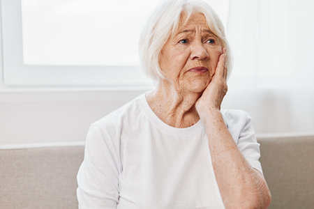 Elderly woman severe tooth pain sitting on the couch, health problems in old age, poor quality of life. Grandmother with gray hair is holding her sore jaw.の写真素材