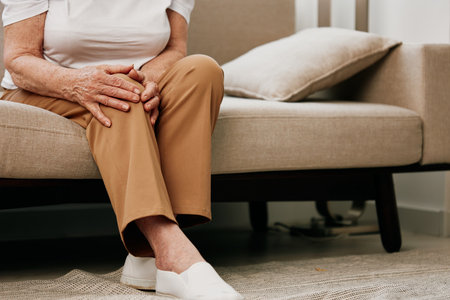 Elderly woman severe pain in her leg sitting on the couch, health problems in old age, poor quality of life. Grandmother with gray hair holds on to her sore knee, problems with joints and ligaments.の写真素材