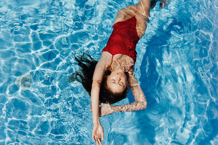 Young woman in the pool in red swimsuit with a beautiful smile lying on the water and swimming in the blue pool in the sun, the concept of relaxing on vacation.の写真素材