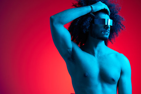 Portrait of fashion man with curly hair on red background with stylish glasses, multinational, colored light, black leather jacket trend, modern concept, sexy body.の写真素材