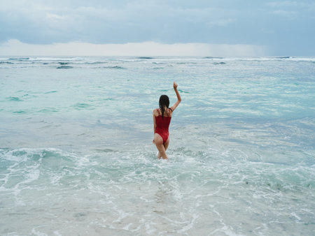 Woman standing with her back to the camera tourist in a red bathing suit sitting on the sand on the beach in the ocean in the waves, travel and beach vacationの写真素材
