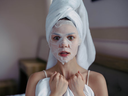 Young woman lying on bed with moisturizing beauty face mask and white towel on her head after shower, lifestyle home skincare, smile.の写真素材