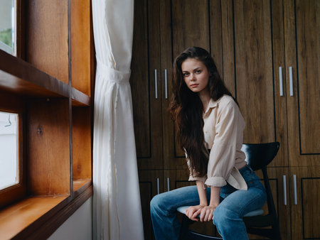 Woman sitting on a chair in home interior near the window, comfort and coziness, lifestyle relaxation on the weekend, pensive look of a beautiful girl.の写真素材