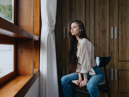 Woman sitting on a chair in home interior near the window, comfort and coziness, lifestyle relaxation on the weekend, pensive look of a beautiful girl.の写真素材