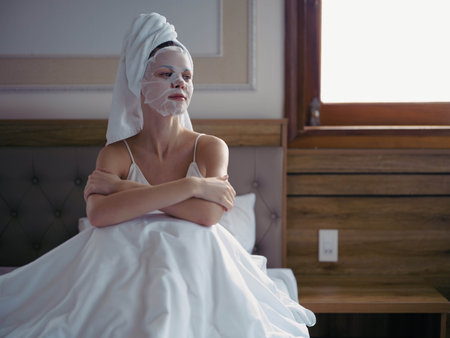 Young woman lying on bed with moisturizing beauty face mask and white towel on her head after shower, lifestyle home skincare, smile.の写真素材